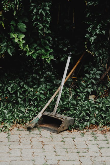 Close-up view of a gardener wearing a white glove and a wristwatch, using a small spade to collect a pile of garden debris, including small branches, leaves, and twigs, into a white compost bag. The g